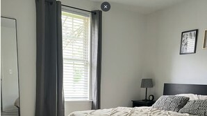 Une chambre à coucher avec un lit, une table de chevet, une lampe, une fenêtre avec des rideaux et un tableau encadré au mur.