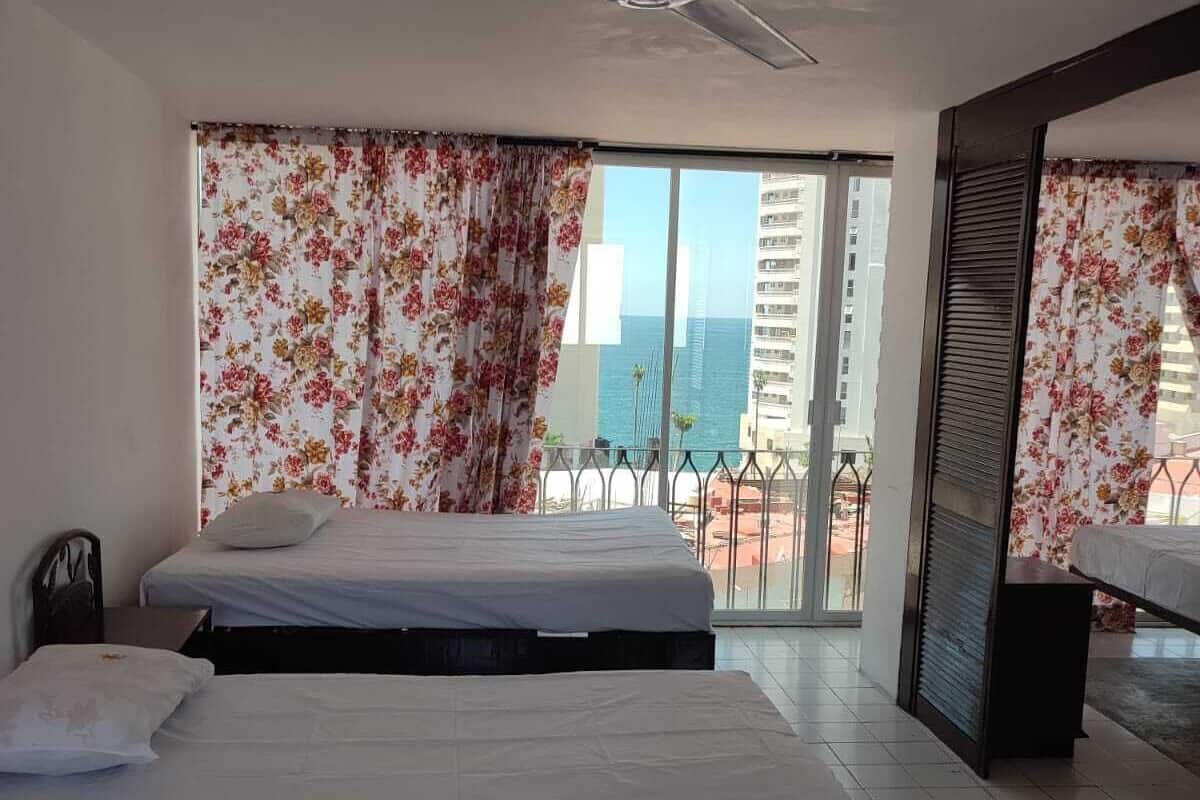 A hotel room with two beds, a ceiling fan, and a balcony with a view of the city and ocean.