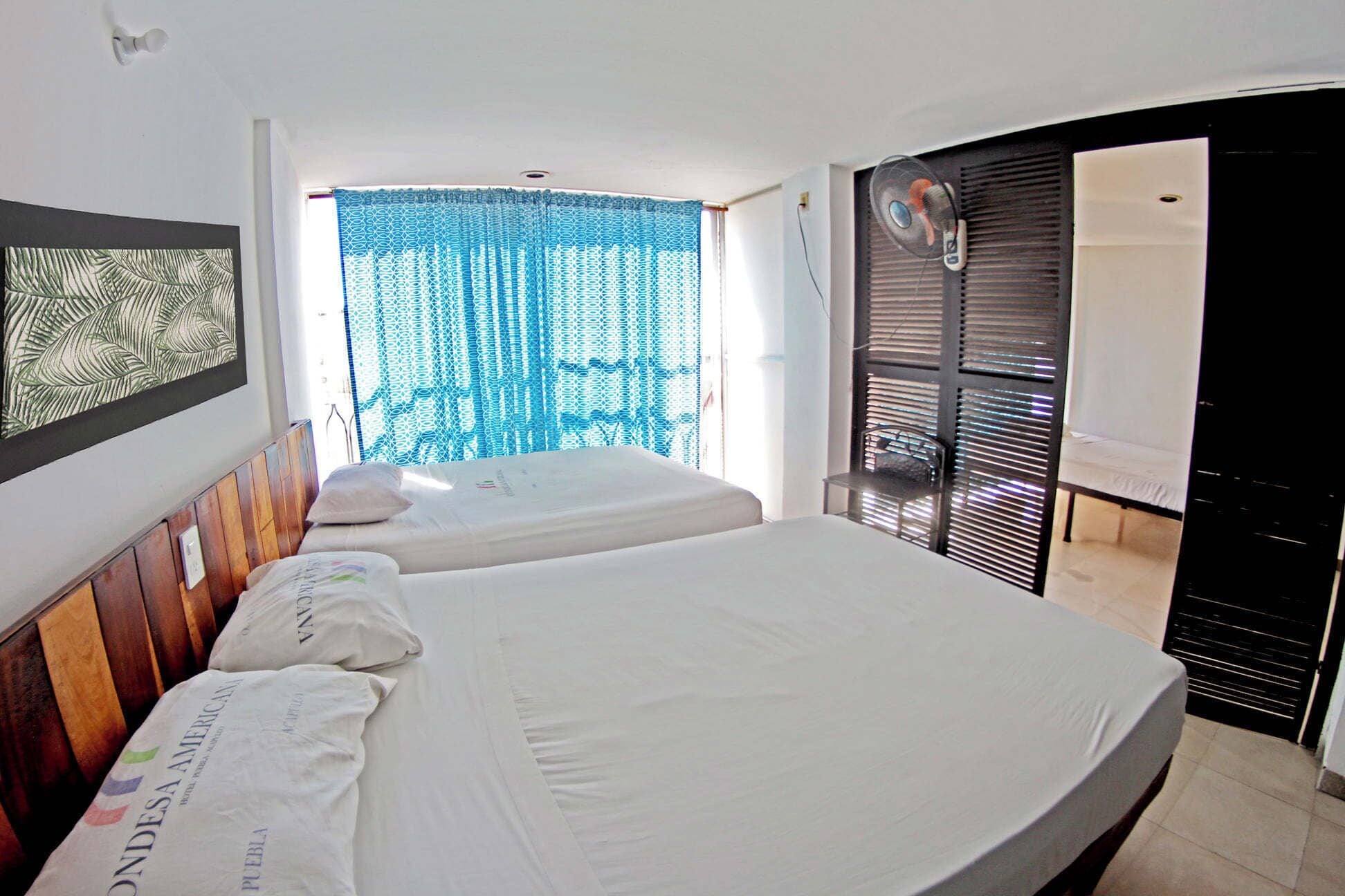 Une chambre d’hôtel avec deux lits, une grande fenêtre avec des rideaux bleus, un ventilateur de plafond et une vue sur l’extérieur.