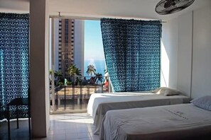A hotel room with two beds, a balcony with a view of palm trees and a building, and a ceiling fan. - Condesa Americana Acapulco (Acapulco)