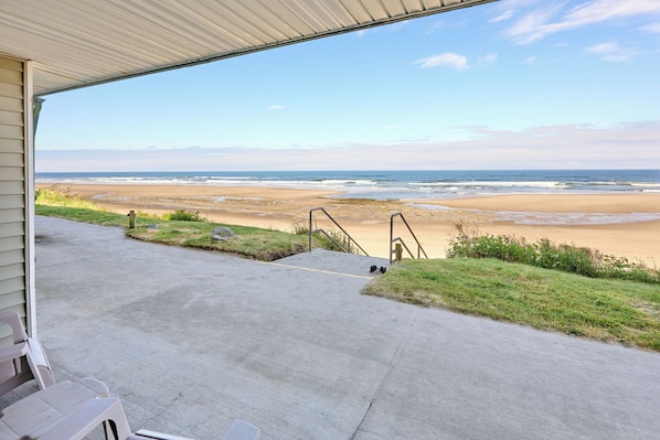Une terrasse couverte, dotée d’un sol en béton et de rampes en métal, qui offre une vue sur une plage de sable fin et les vagues de l’océan.