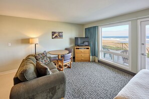 A hotel room with a sofa, a small round table, a TV on a wooden stand, and a large window with a view of the beach.