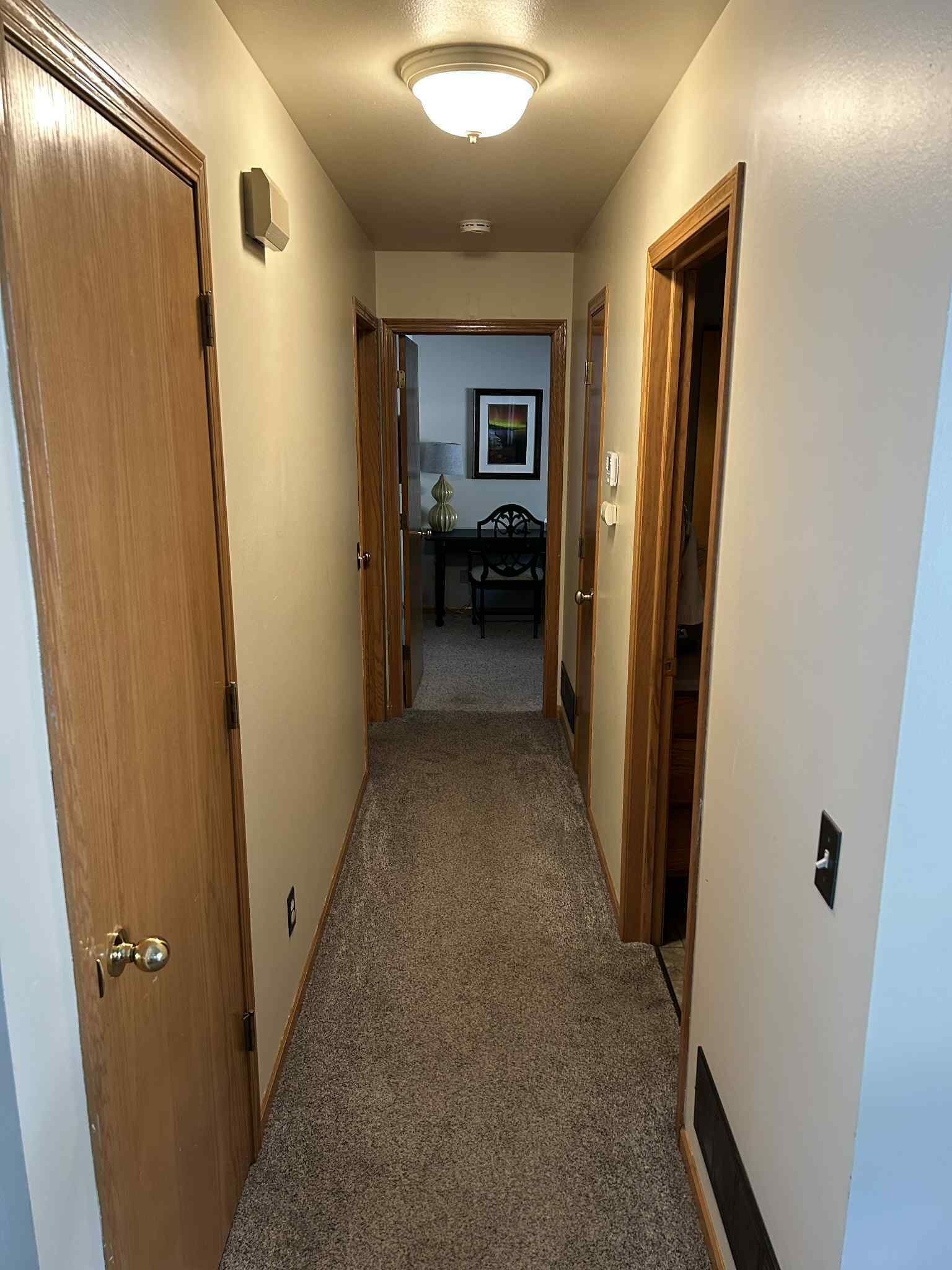 A narrow carpeted hallway with wooden doors on either side, leading to a room with a bed and a lamp.