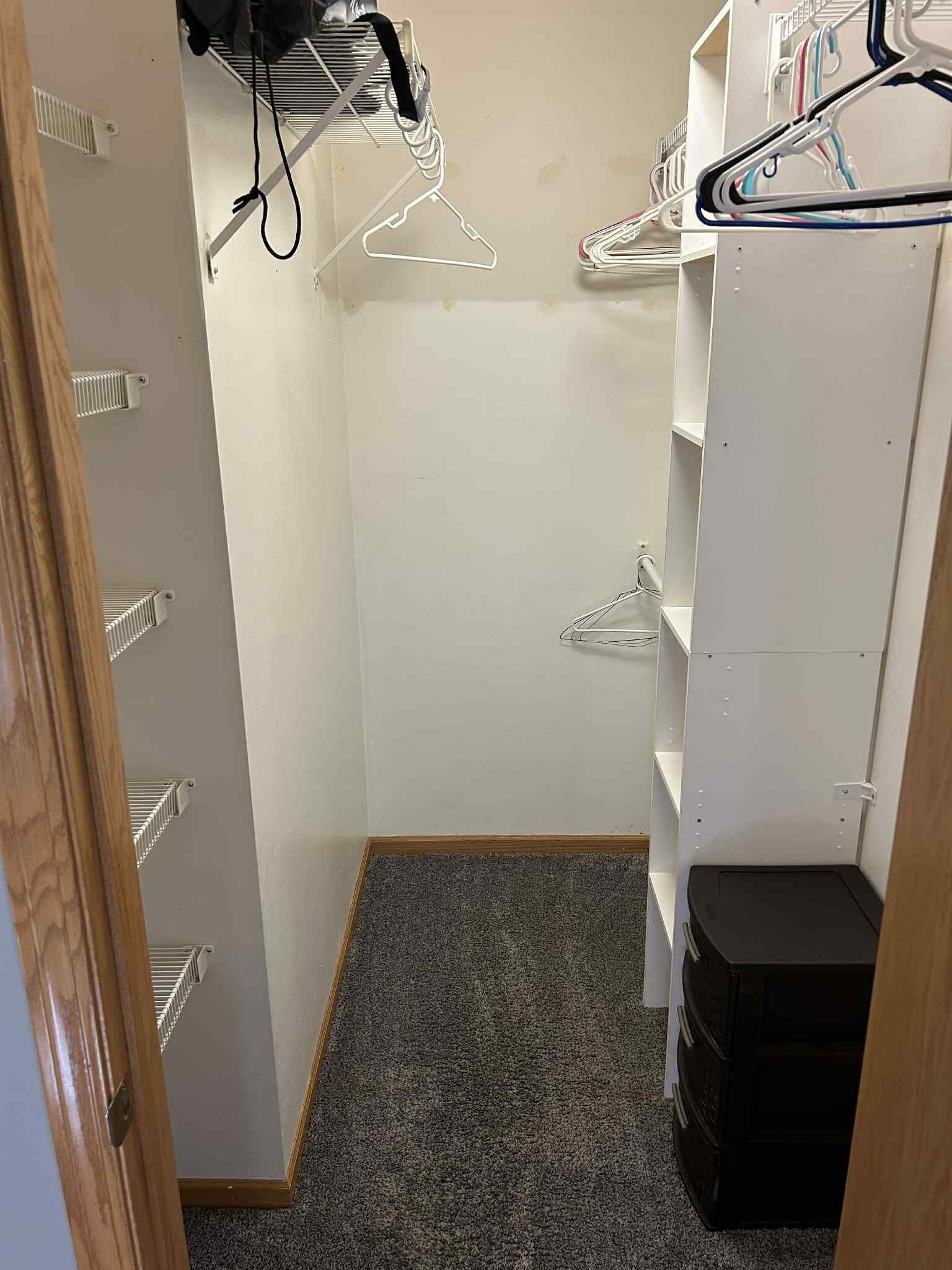A walk-in closet with white shelving units, a black storage bin, and a hanging rack with clothes.