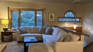 Una sala de estar acogedora con un sofá modular, una mesita de centro y una lámpara. Amplias ventanas ofrecen luz natural y vistas a árboles.