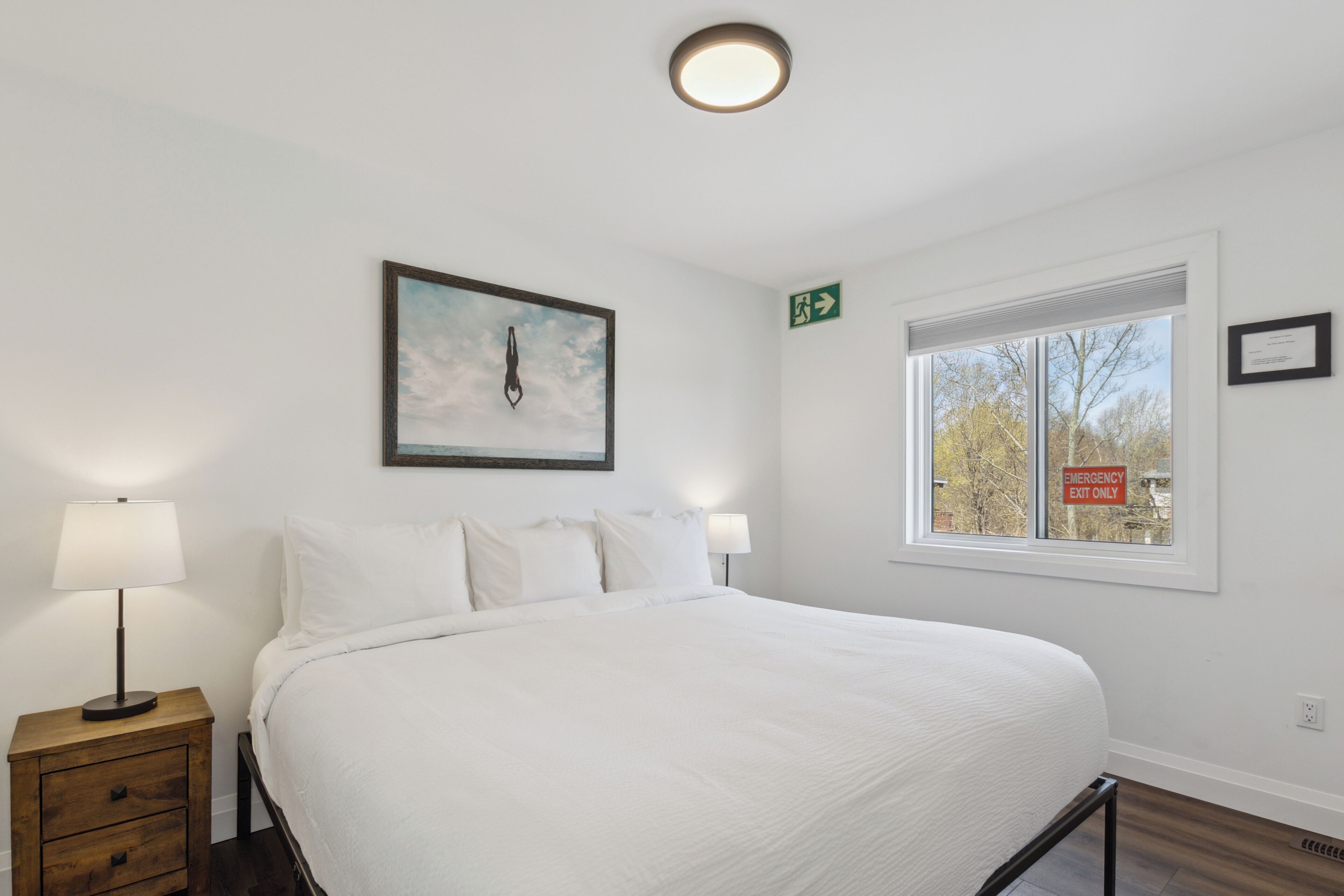 Un lit bien fait, avec du linge de lit blanc, une table de chevet avec une lampe, un tableau encadré au mur et une fenêtre avec un panneau indiquant la sortie de secours.