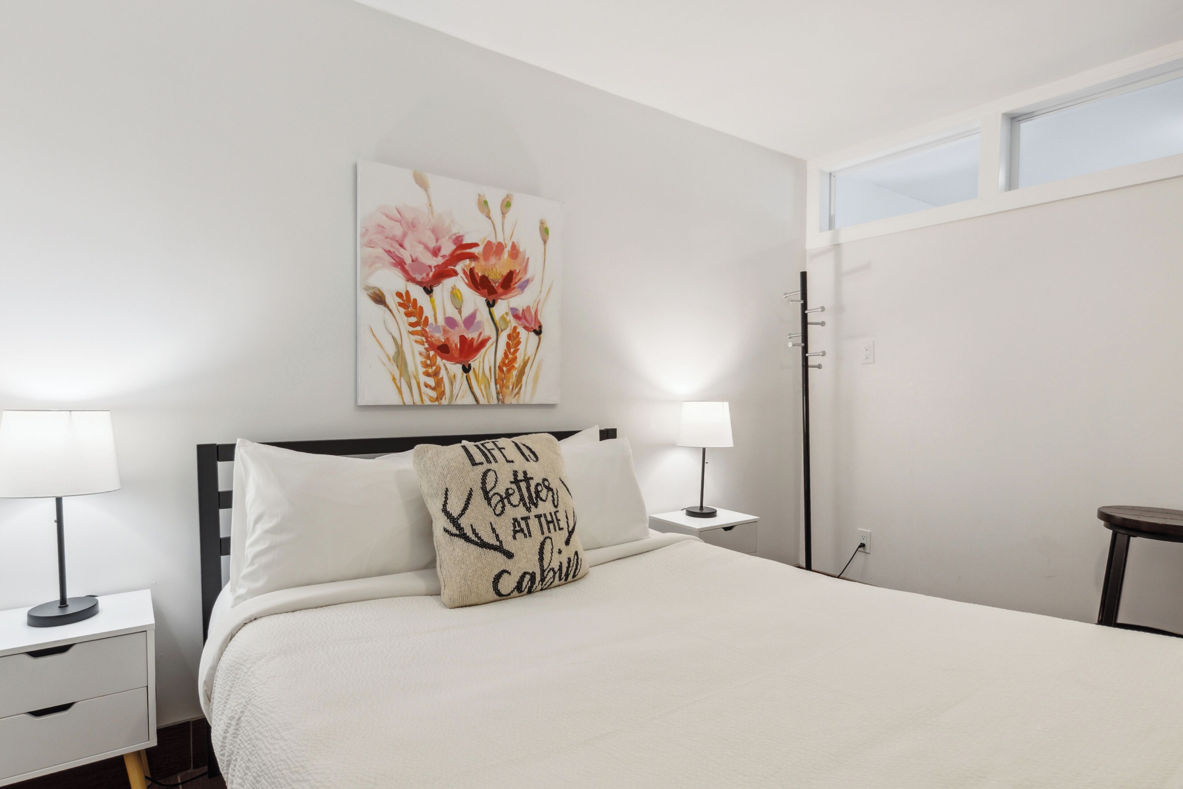 Une chambre à coucher bien rangée, avec un lit, deux tables de chevet, une commode, une fenêtre et une œuvre d’art accrochée au mur.