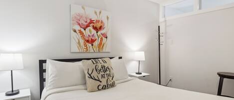 Une chambre à coucher bien rangée, avec un lit, deux tables de chevet, une commode, une fenêtre et une œuvre d’art accrochée au mur.