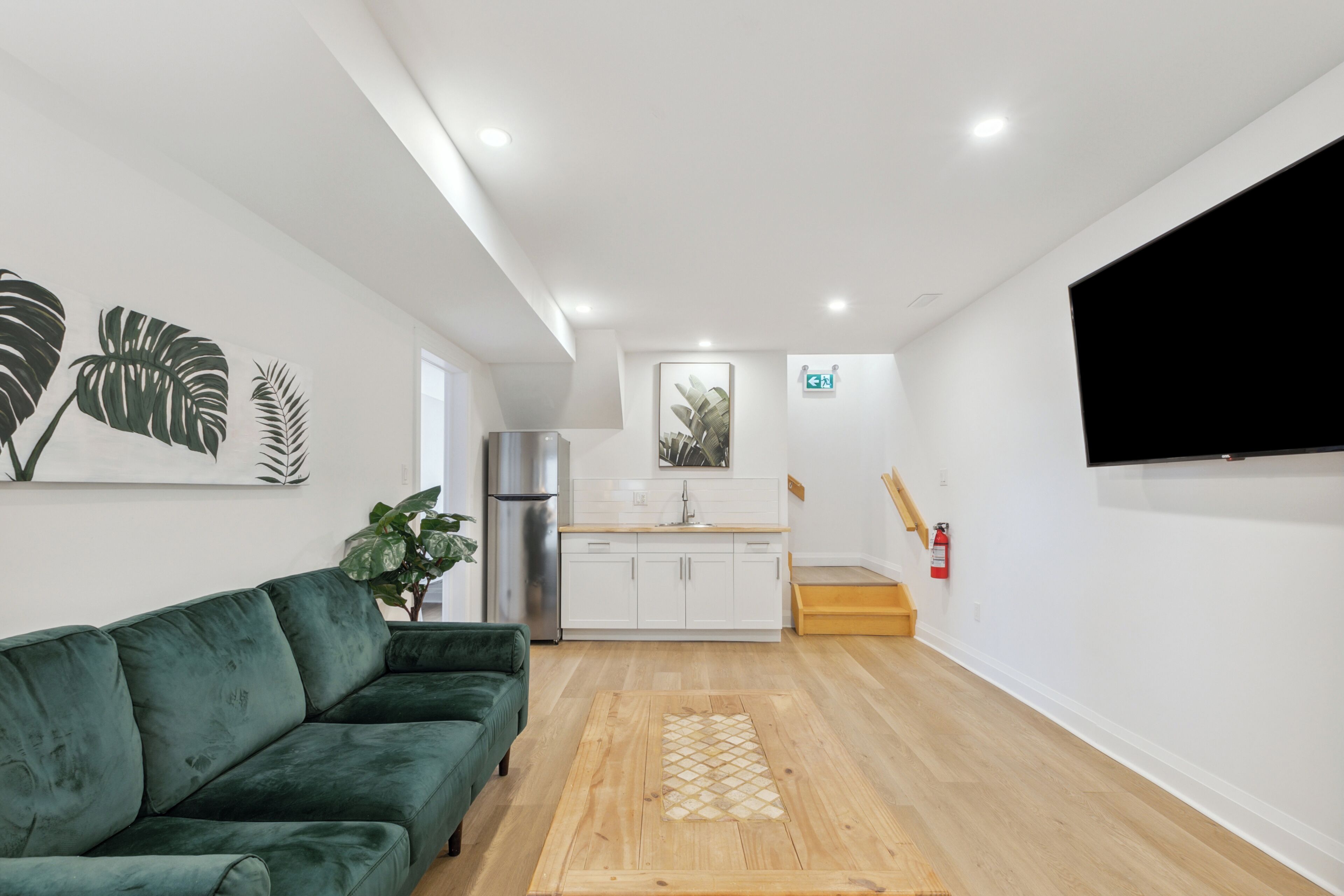A modern living room with a green sofa, wooden flooring, a kitchen area in the background, and a mounted TV on the wall.