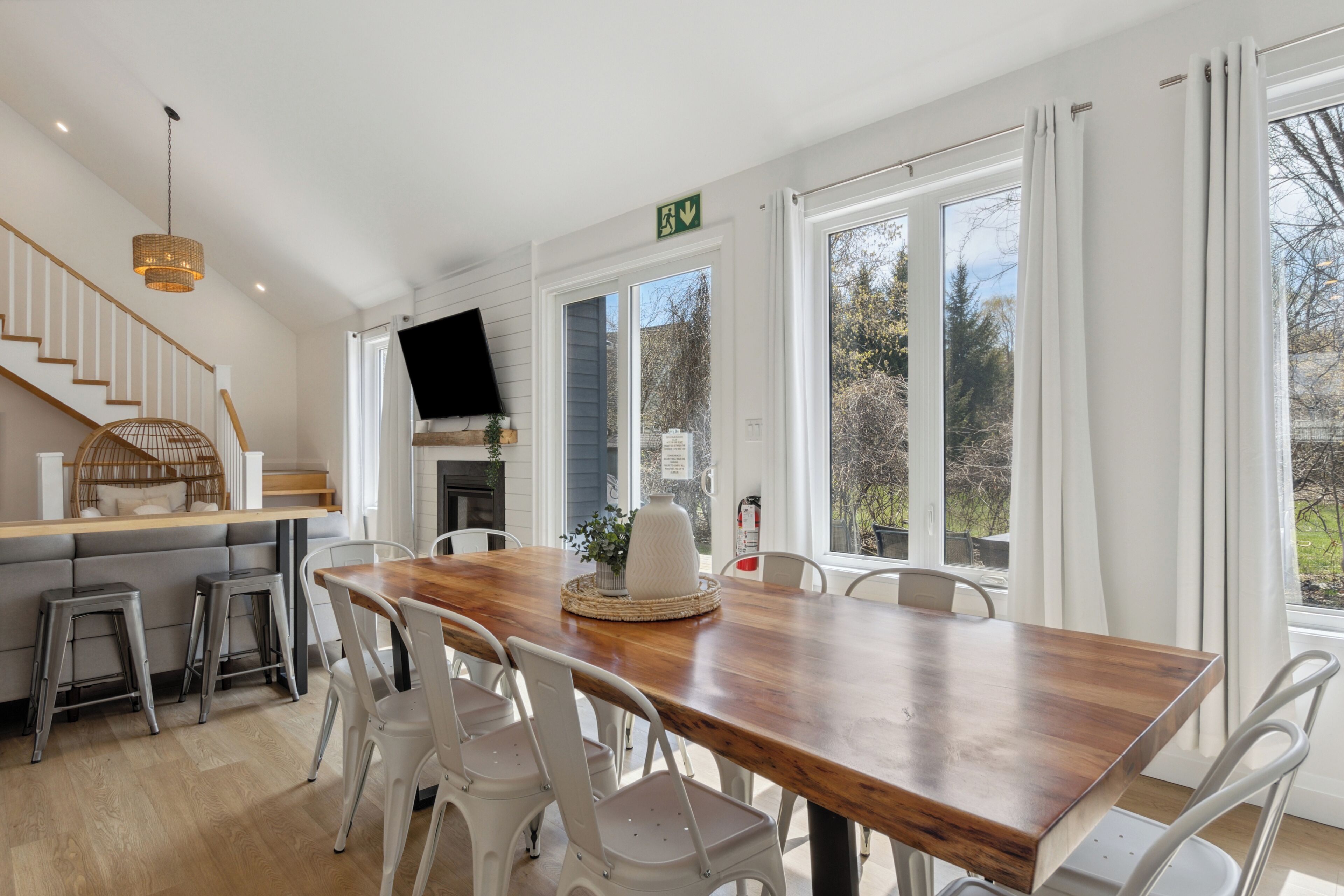 A spacious dining area with a wooden table and white chairs, a staircase with a woven light fixture, and large windows offering a view of the outdoors.