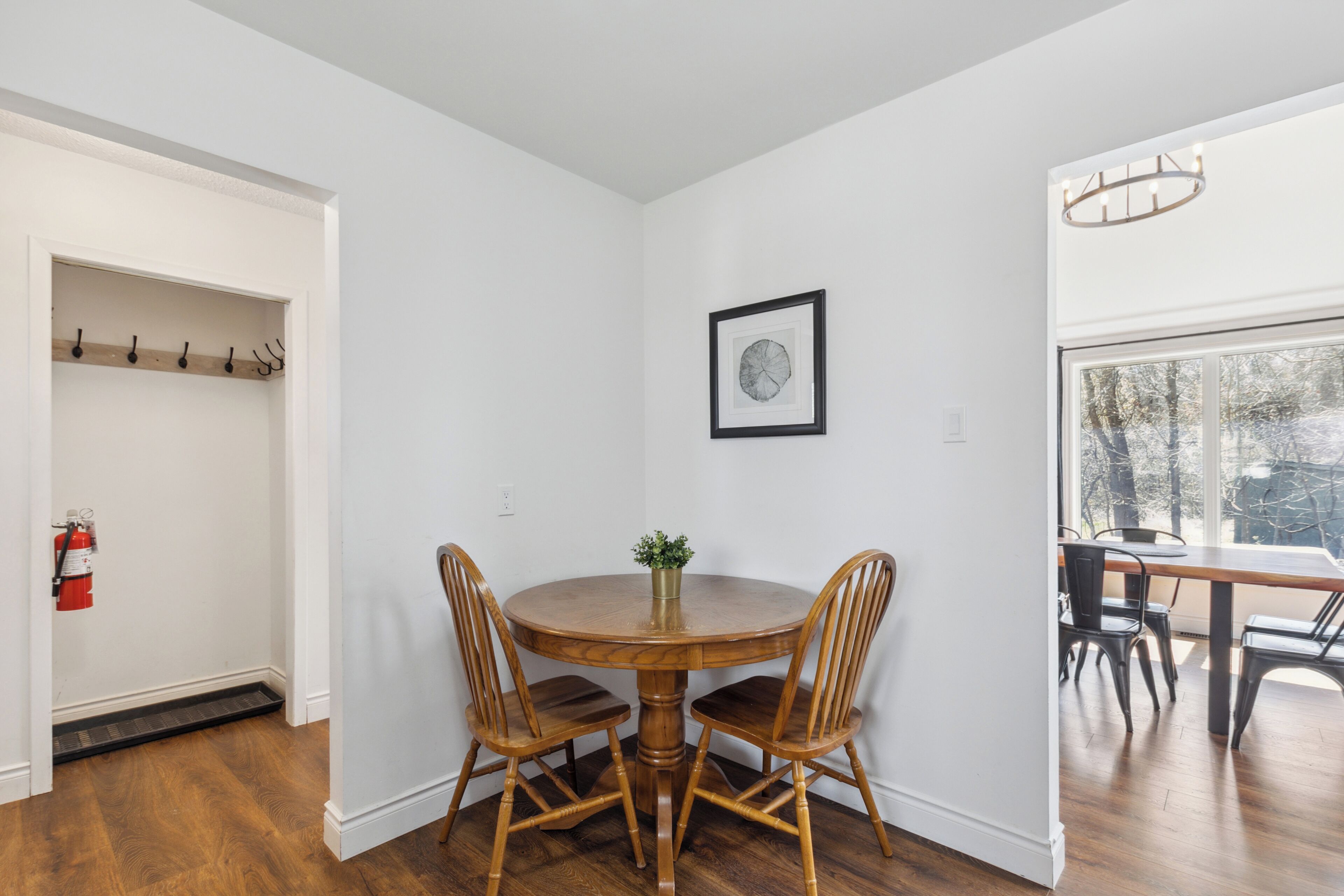A dining area with a round wooden table and four chairs, a framed picture on the wall, and a view through a window.