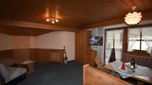 L’intérieur chaleureux d’un chalet avec un plafond en bois, un téléviseur fixé au mur, une table à manger surmontée d’un abat-jour conique rouge et une fenêtre ornée de rideaux légers.