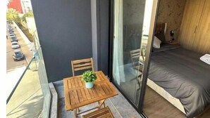 Ein Balkon mit Holztisch und Stühlen, einem kleinen Topfpflanzen und einer Glastür, die zu einem Schlafzimmer mit einem Bett führt.