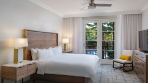 Ein Hotelzimmer mit einem großen Bett, einem Fernseher, einem Schreibtisch mit Lampe, einem Sessel und einem Balkon mit Blick auf Palmen.