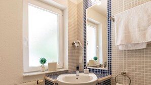 A bathroom with a white sink, a mirror, a towel rack, and a window with blinds. - Memory 04 - Double room (approx. 25 m²) with balcony on the 1st floor (Bischofsheim in der Rhön)