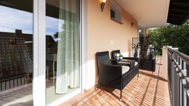 A balcony with a sofa, chairs, and a small table, overlooking a garden and buildings. - Memory 04 - Double room (approx. 25 m²) with balcony on the 1st floor (Bischofsheim in der Rhön)