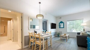 A modern living room with a dining area, a TV mounted on the wall, and a window with blinds. - Inspiration condo near Disney with free shuttle (Orlando)