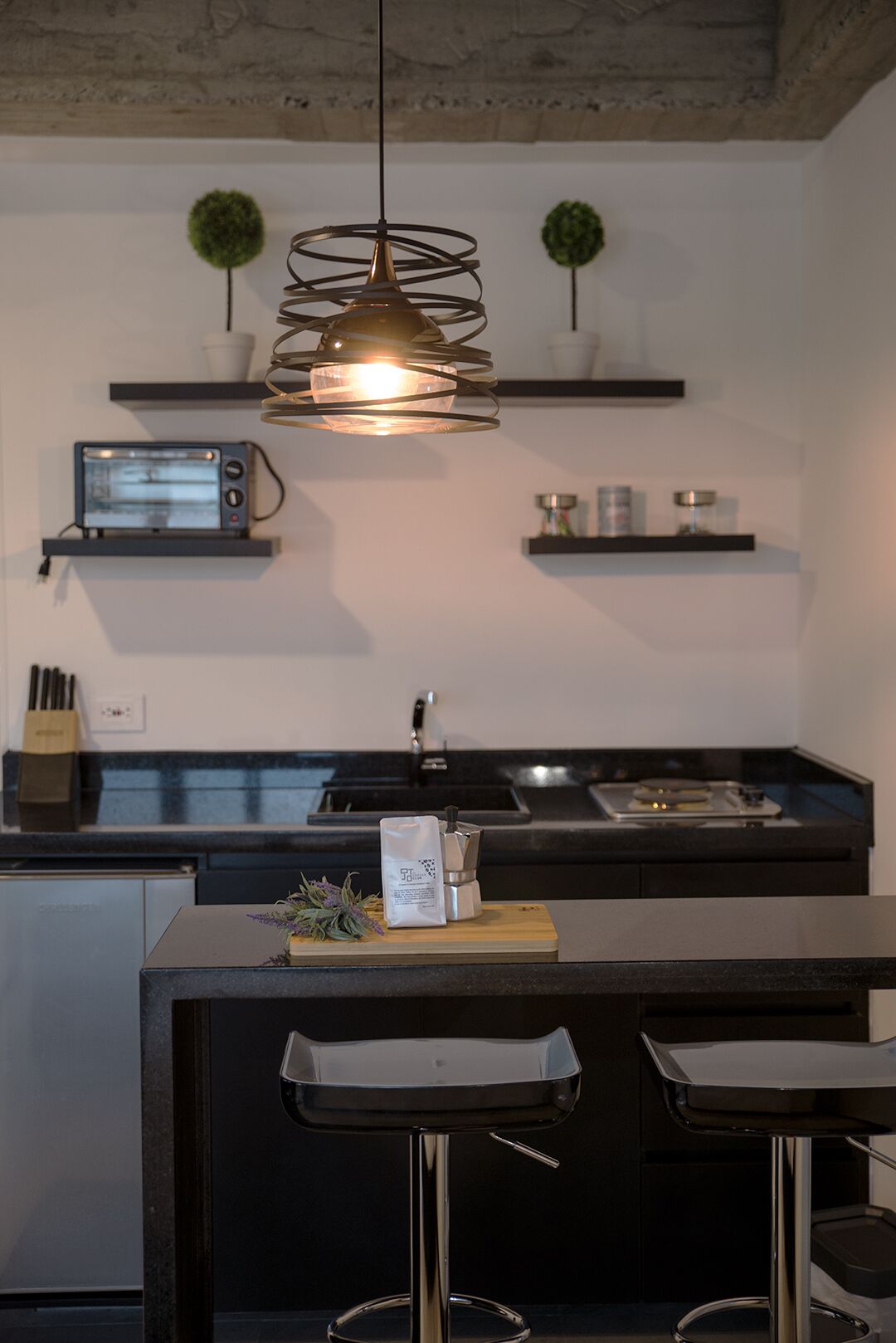 A modern kitchen with a central island, black bar stools, a hanging pendant light, and a toaster oven on a shelf.