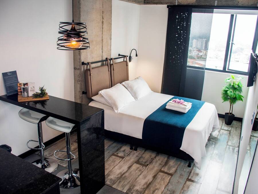 Una habitación de hotel moderna con una cama, un escritorio y un taburete de bar. Se ve una ventana con vistas a edificios.