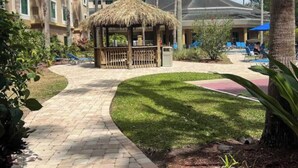A paved pathway leading to a thatched gazebo surrounded by palm trees and a pool area with lounge chairs. - Renovated Condo near Disney Free Shuttle to Parks (Orlando)