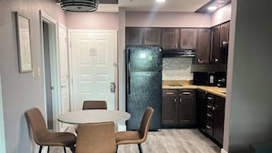 A modern kitchen with dark wooden cabinets, a black refrigerator, and a microwave. There is a dining area with a round table and four chairs. The floor is covered with light wood laminate. - Renovated Condo near Disney Free Shuttle to Parks (Orlando)