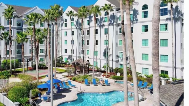 A hotel with a swimming pool surrounded by palm trees and blue lounge chairs. - Condo near Disney-free shuttle to all theme parks (Orlando)