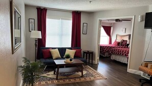 A living room with a dark-colored sofa, a coffee table, a potted plant, and a patterned rug. There is a bed with red bedding in the background.