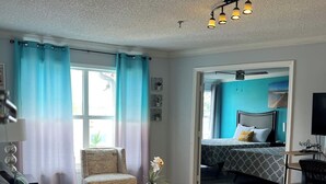 A modern living room with a sofa, a round coffee table, and a small potted plant. There is a bed with a blue headboard in the background, and a ceiling fan above. - Condo Near Disney-Universal-Epic-Sea World (Kissimmee)