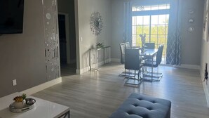 A modern living room with a dining area, light wood flooring, a flat-screen TV, and a large window with curtains. - Amazing Condo near Disney World (Kissimmee)