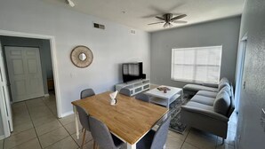 Una sala de estar moderna con comedor. Incluye una mesa de comedor de madera con sillas, un sofá gris, un televisor y un ventilador de techo.
