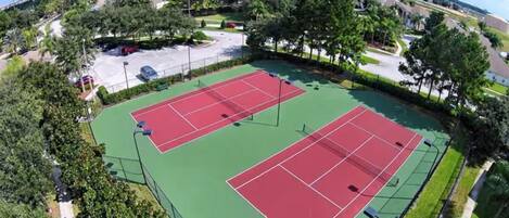 An aerial view of a tennis court complex with multiple courts, surrounded by greenery and a parking area.