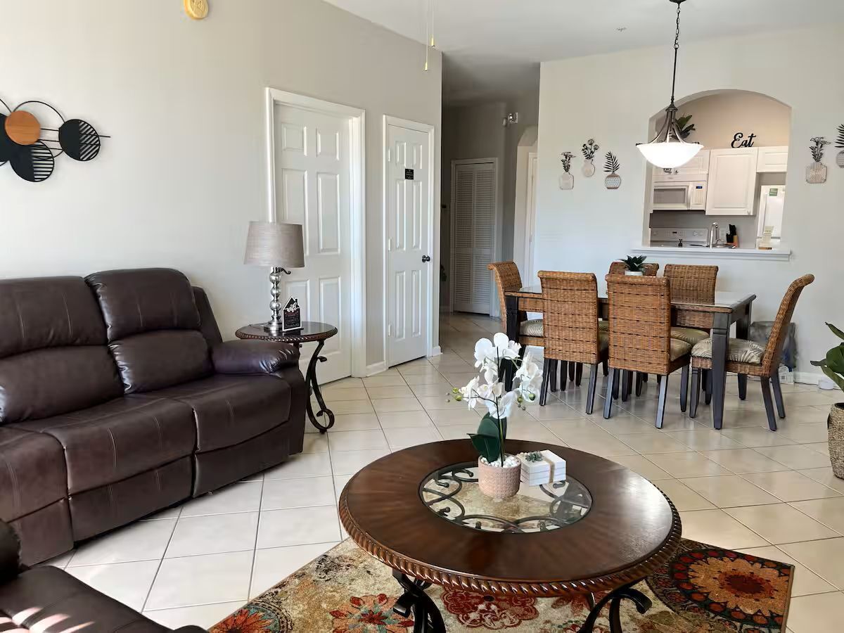 A living room with a brown leather sofa, a round coffee table with a decorative centerpiece, a dining area with wicker chairs, and a kitchen in the background.