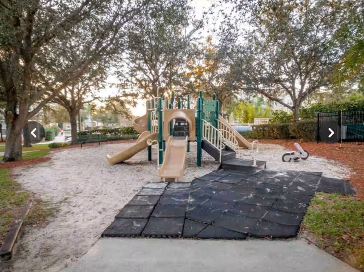 A playground with slides and climbing structures, surrounded by trees and a paved area.