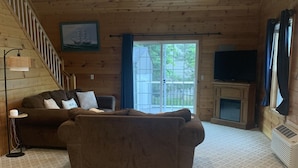 A cozy living room with a brown sofa, a television, a fireplace, and a staircase leading to a balcony. - Limestone Landing - Pool, firepit, covered patio, & more! (Kelleys Island)