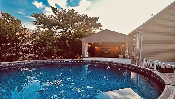 A swimming pool with a covered patio area, surrounded by trees and a building. - Limestone Landing - Pool, firepit, covered patio, & more! (Kelleys Island)