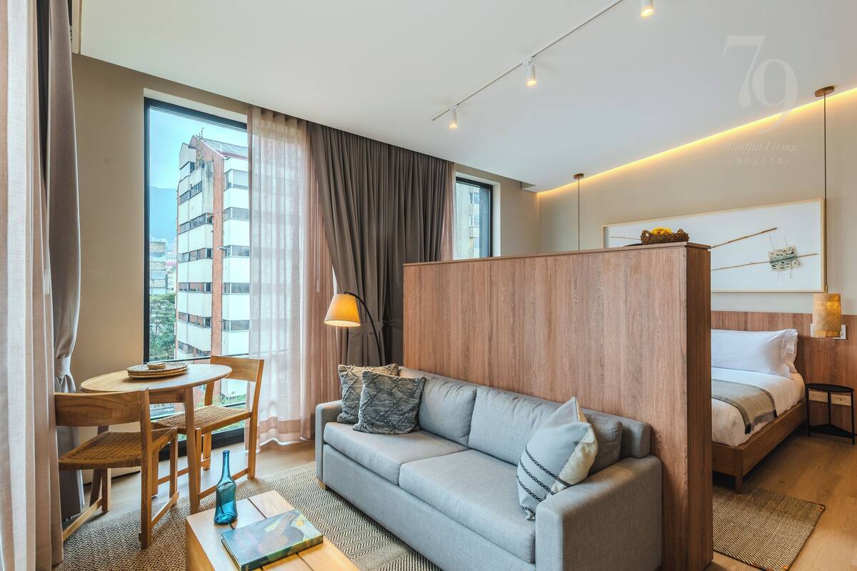 A modern hotel room with a sofa, a small dining table, a bed, and a view of a building outside.