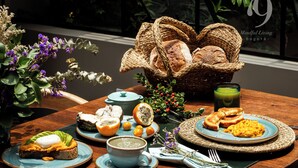 Una mesa de madera dispuesta con alimentos para el desayuno, incluyendo huevos, tostadas y fruta, junto con cestas tejidas y un vaso de vidrio verde.