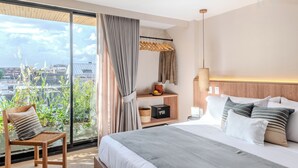 Una habitación de hotel moderna con una cama grande, una silla de madera, una mesita de noche, una estantería con objetos decorativos y vistas a la ciudad a través de grandes ventanales.