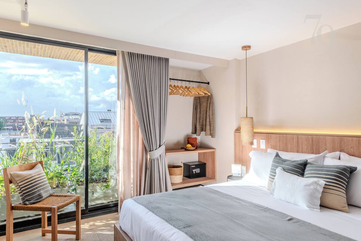 A modern hotel room with a large bed, a wooden chair, a bedside table, a shelf with decorative items, and a view of the city through large windows.