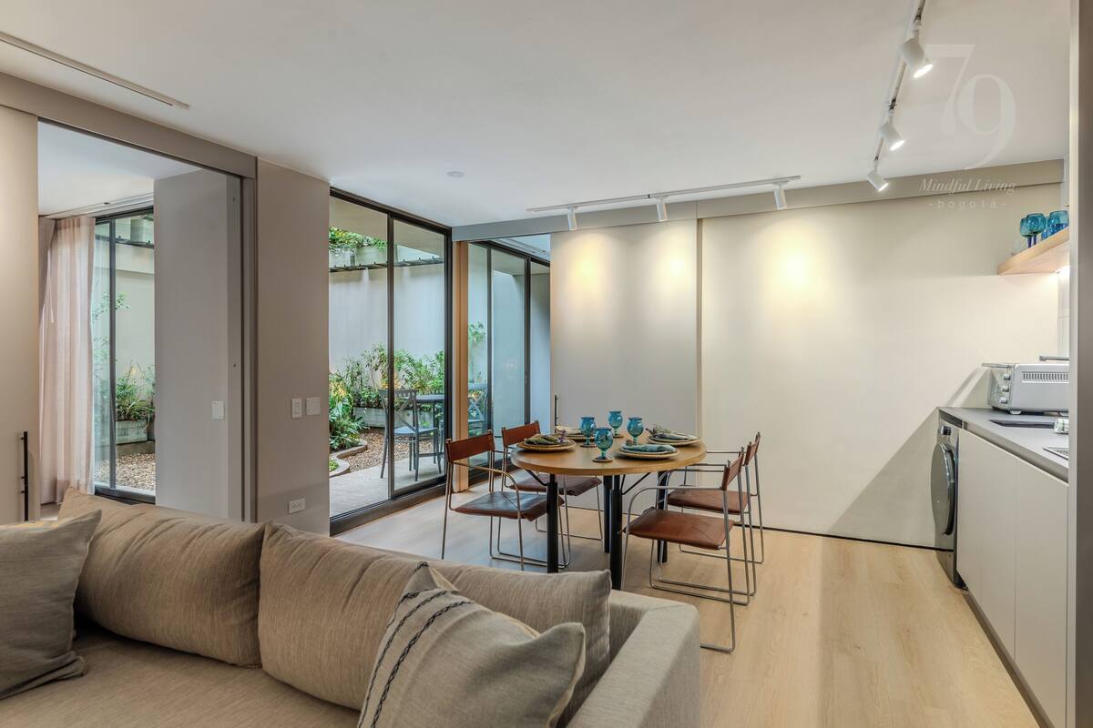 A modern living room with a sofa, a dining table with chairs, and a kitchen area with a washing machine.