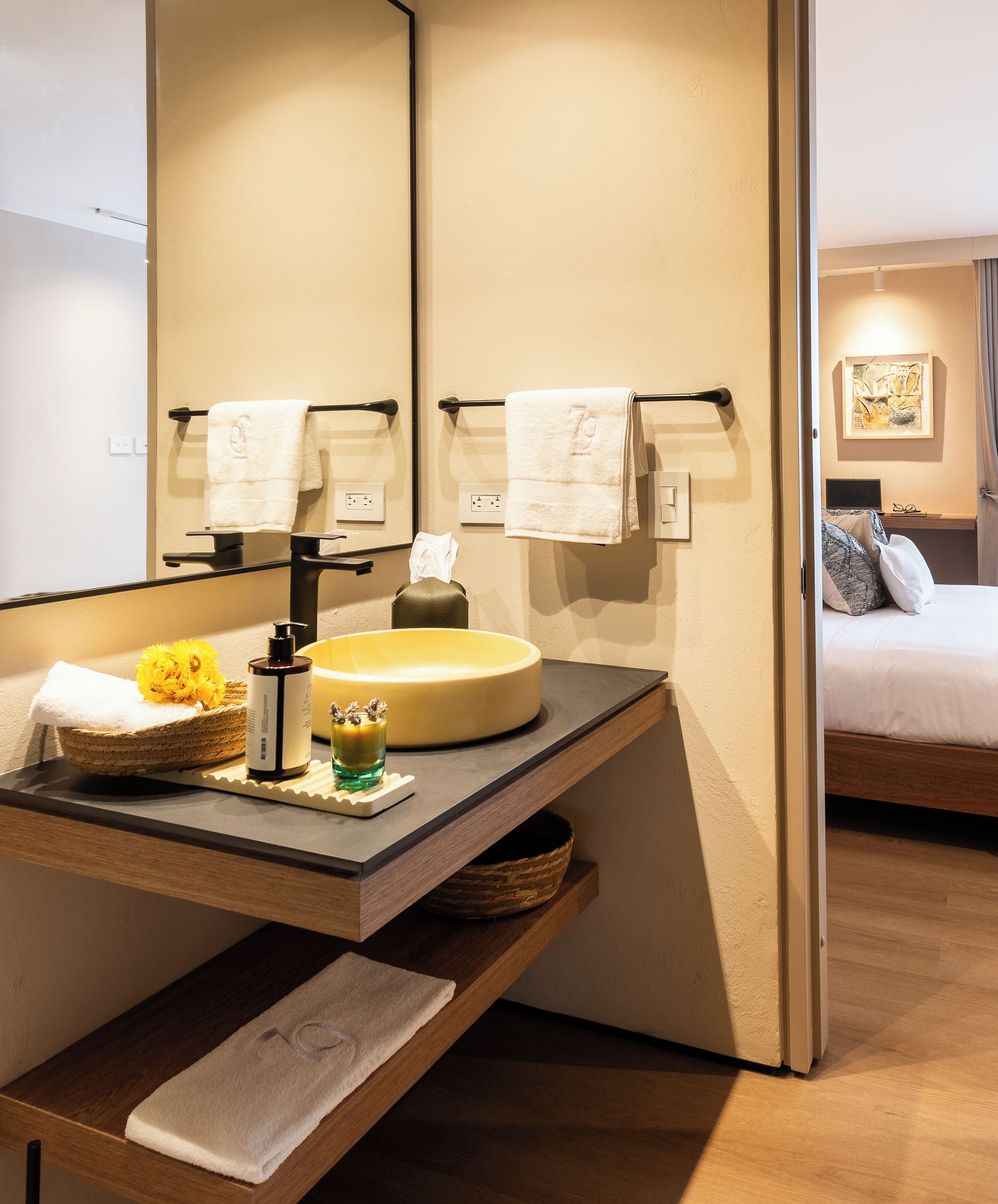 A modern bathroom with a large mirror, a round sink, and a bedside table with a towel.