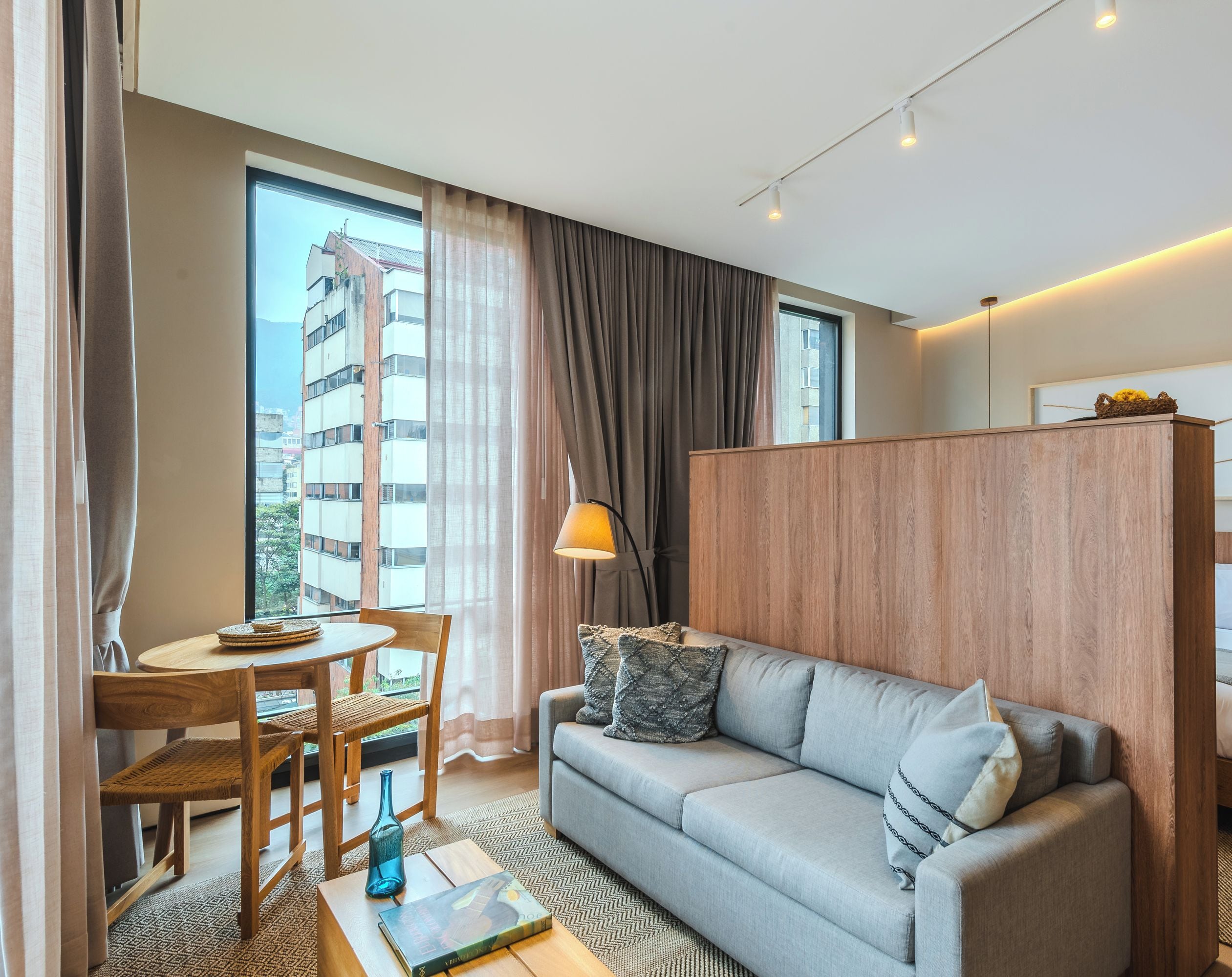 A modern hotel room with a sofa, a small dining table, a bed, and a view of a building outside.