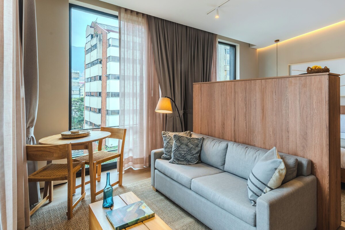 Habitación de hotel moderna con sofá, una pequeña mesa de comedor, cama y vistas a un edificio.