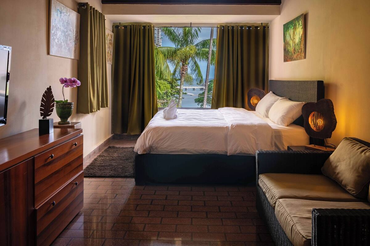 Una camera d'albergo con un letto grande, una poltrona, una cassettiera e vista su palme e un ponte.