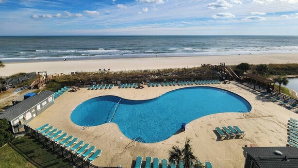A beachside pool with a curved design, surrounded by lounge chairs and a wooden deck. - Oceanfront Gem (Myrtle Beach)