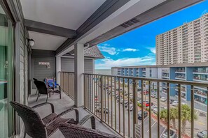 A balcony with wicker chairs, a small table, and a view of a parking lot and buildings.