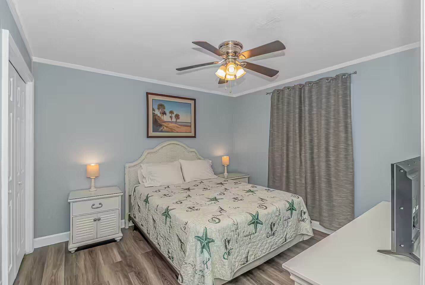 Un dormitorio con cama, mesitas de noche, ventilador de techo, un cuadro y una cortina.