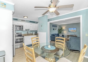 A modern kitchen with stainless steel appliances, a dining area with a glass table and wooden chairs, and a living room with a blue sofa and a television.