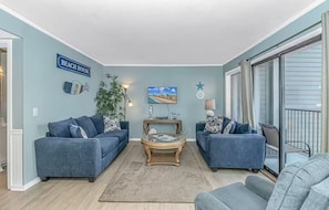 A living room with two blue sofas, a coffee table, and a TV. There is a sign on the wall that says "BEACH HOUSE".