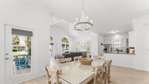 Une cuisine spacieuse avec des meubles blancs, un coin repas doté d’une table en verre, et une vue sur un jardin avec des chaises Adirondack bleues.
